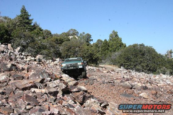1991 Ford Bronco navigating a steep, rocky trail with trees and blue sky in the background
