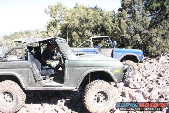Two Ford Broncos on a rocky trail, one green and one blue. The green Bronco is in the foreground with a driver