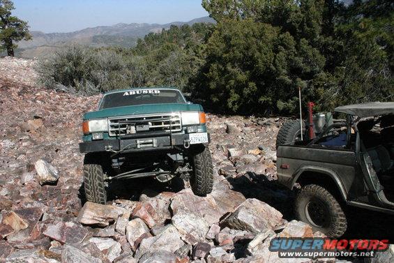 Green Ford Bronco with 'ABUSED' sticker on windshield navigating rocks, another Bronco to the right