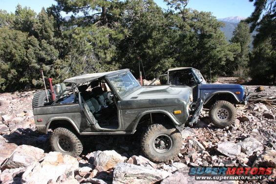 Two Ford Broncos parked on a rocky trail with trees and mountains in the background. The front Bronco is dark green and open-topped; the rear is blue