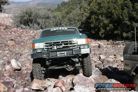 Green SUV with 'ABUSED' sticker on windshield, driving over rocks. Another vehicle is partially visible to the right