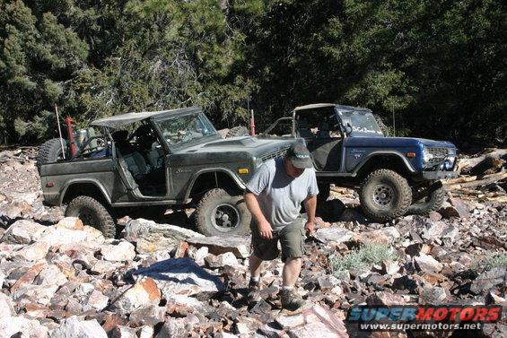 Two Ford Broncos navigating a rocky, wooded trail. The front Bronco is dark green and open-topped; the rear is blue. A man walks between them