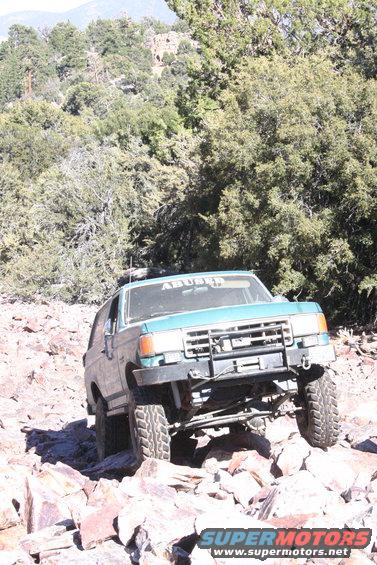 Teal Ford Bronco climbing over rocks on a trail. Text on windshield reads 'ABUSED'