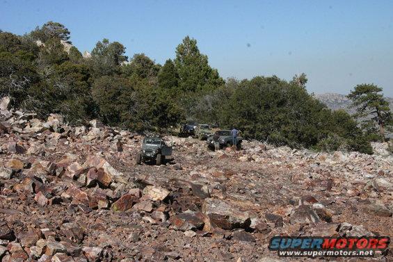 Four-wheel drive vehicles navigate a rocky, wooded hillside under a clear blue sky