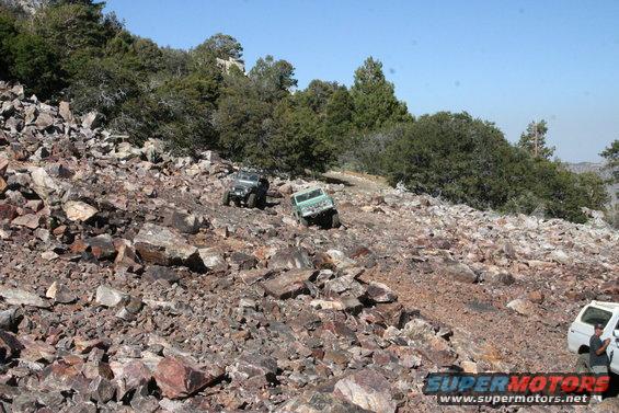 Two off-road vehicles navigate a rocky, wooded hillside under a clear blue sky