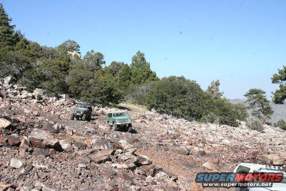 Two off-road vehicles navigate a rocky, mountainous trail under a clear blue sky