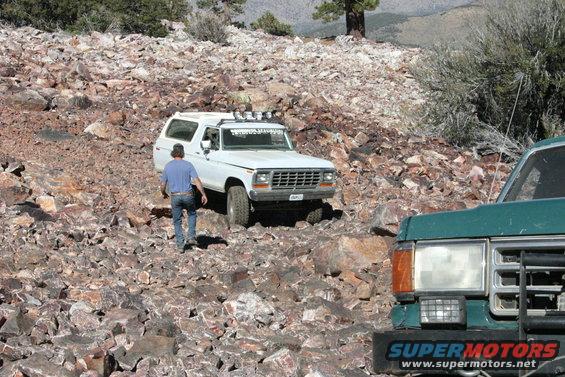 White Ford Bronco driving on a rocky trail, with a green truck in the foreground. A person is walking near the Bronco