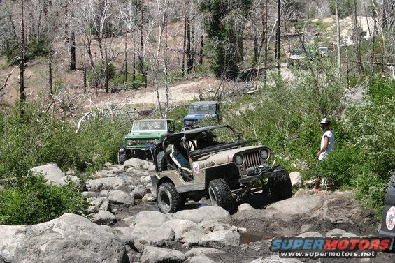 Several Jeeps navigate a challenging rocky off-road trail with trees and brush on either side