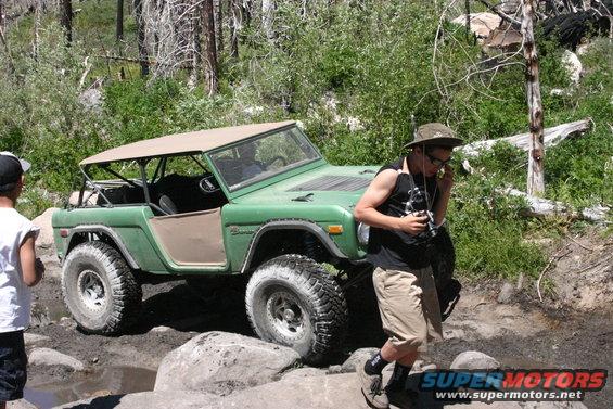 Green first-generation Ford Bronco with tan top, off-roading on rocks