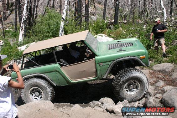 Green Ford Bronco with tan soft top navigating rocky terrain. A person is taking a photo of the vehicle