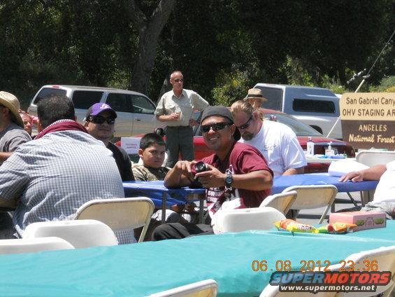 People gathered at tables outdoors with vehicles in the background