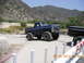 1986 Ford Bronco - A lifted blue pickup truck tows a black pickup truck on a road with a mountainous backdrop.