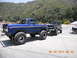 1986 Ford Bronco - A lifted blue Ford Bronco tows a black Ford pickup truck on a sunny day with a mountainous backdrop.