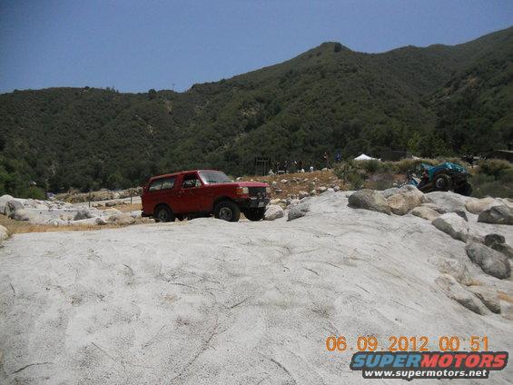 Red 1986 Ford Bronco off-roading on rocky terrain with a blue ATV in the background