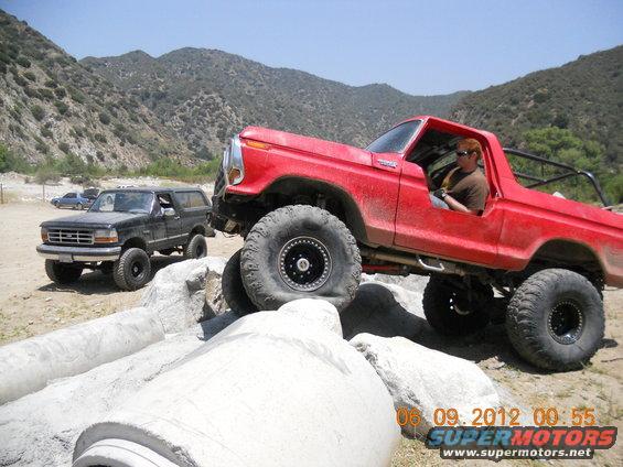 Red off-road Bronco climbing over rocks, black Bronco behind it