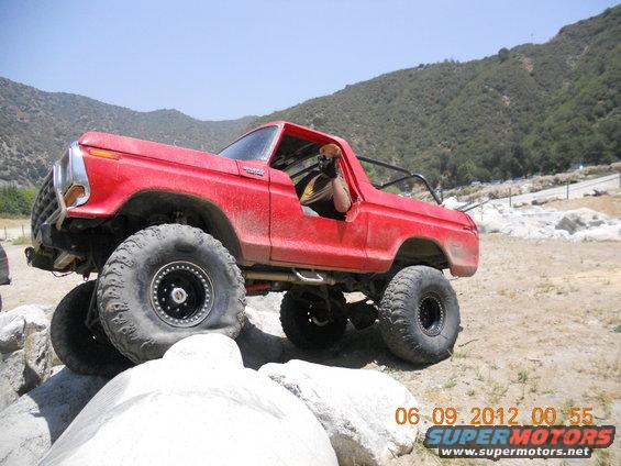 Red 1978 Ford Bronco with large off-road tires climbing over rocks. Driver is visible in the open door