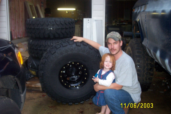 Man and young child posing with a stack of large off-road tires next to a dark-colored vehicle