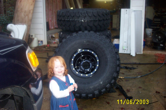 Child in blue dress stands before stacked off-road tires in a garage