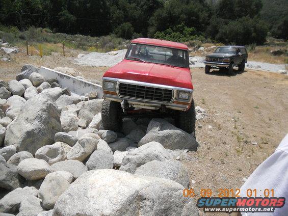 Red 1978 Ford Bronco navigating large rocks on a trail; another Bronco is visible in the background