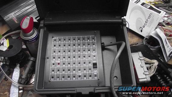 Open black box with a grid of numbered fuse slots, some labeled. Wires and a manual are nearby