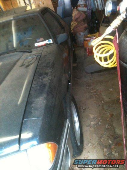 Front corner of a dark 1990 Ford Mustang parked in a garage. The car has a dusty finish and aftermarket wheels