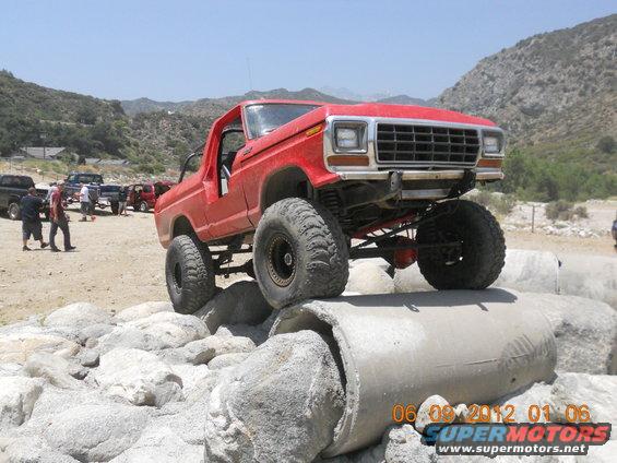 Red lifted Ford Bronco with large tires on concrete pipes, other vehicles and people in background