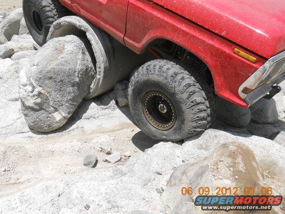 Red off-road vehicle with large tires climbing over rocks and a concrete pipe