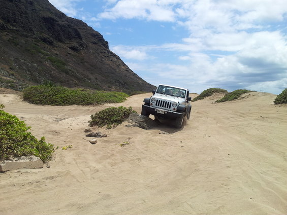 Silver Jeep Wrangler driving on a sandy, rocky trail with a large mountain in the background