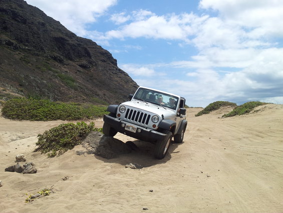 White Jeep Wrangler driving off-road on sand and rocks, with a mountain and cloudy sky
