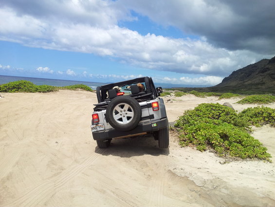 silver Jeep Wrangler with spare tire on back driving on sand near ocean
