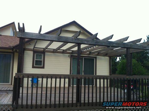 Backyard deck with wooden pergola, beige house with brown trim, and a blue lawn chair