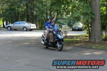 Two people on a blue scooter with two cars in the background