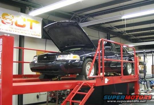 Dark blue Ford Crown Victoria on a red car lift with hood open. Supermotors.net logo visible
