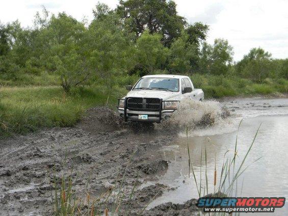 White pickup truck splashing through mud and water, trees and brush in background