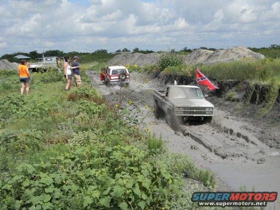 Silver pickup truck driving through mud, another SUV behind it, people watching