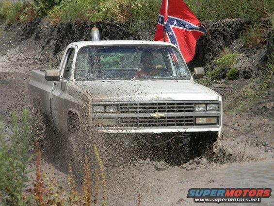 White pickup truck driving through mud with a confederate flag flying behind it