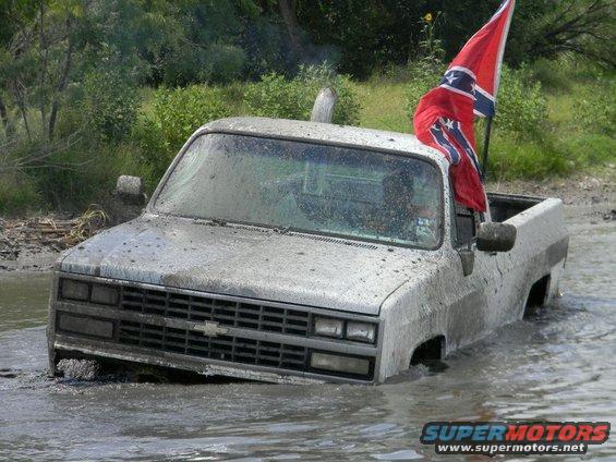 Mud-covered pickup truck stuck in water, Confederate flag flying from the bed. Grille indicates 1985 model