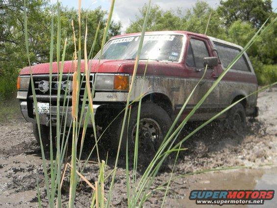 Red and tan Ford Bronco driving through mud, front 3/4 view, covered in mud
