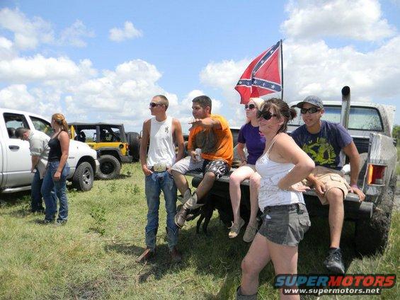 Group of people with a confederate flag and several off-road vehicles in a muddy field