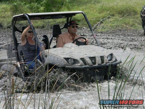 Two people driving a muddy side-by-side through water and reeds