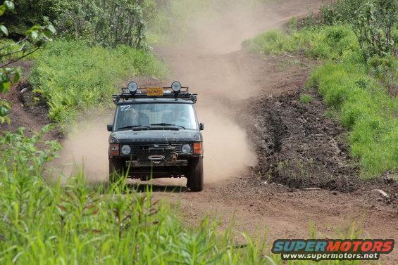 Black Range Rover driving on a dirt road, kicking up dust. It has a roof rack with lights and a winch on the front