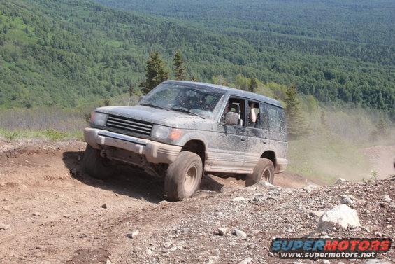 Gray Mitsubishi Pajero off-roading on a dirt hill, front-left tire angled up, dust trailing