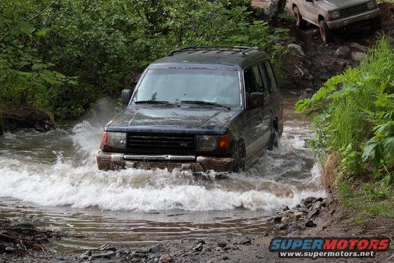 Dark SUV driving through water, another SUV on a muddy incline behind it