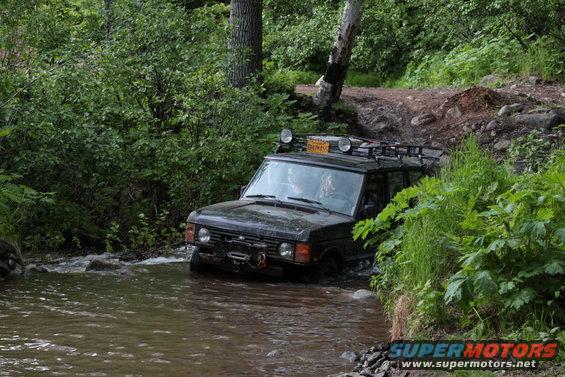 Black Range Rover Classic driving through a shallow stream with a roof rack and auxiliary lights