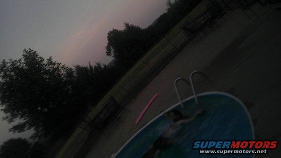 Swimming pool with ladder and pink pool noodle, trees and fence in background at dusk. Supermotors.net logo in corner