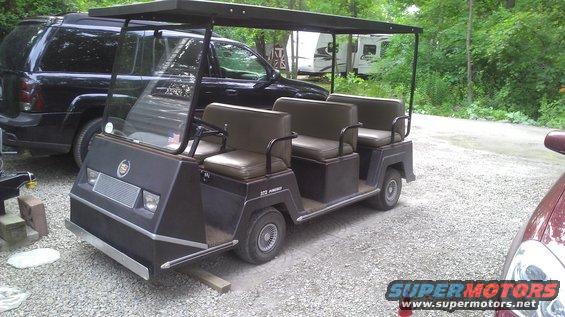 Black six-seater golf cart with a canopy, parked next to a black SUV and a red car