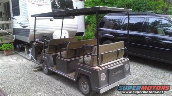 Dark golf cart with Cadillac badging, tan seats, and a canopy, parked next to a black SUV and a trailer