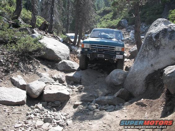 Green SUV with winch on a rocky trail, front view. Another white SUV is visible in the background