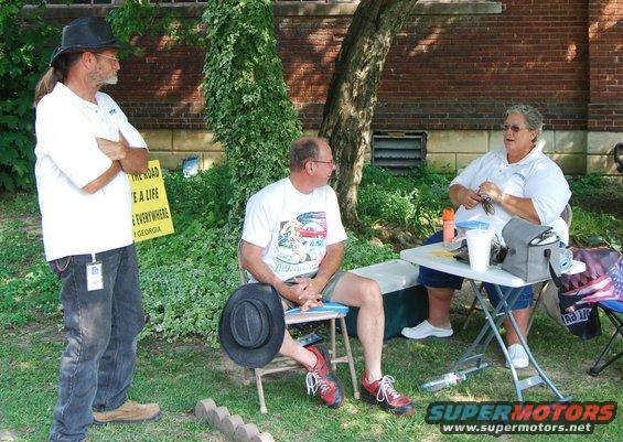 Three people are gathered outdoors, two sitting at a table and one standing. A yellow sign is visible behind the standing person