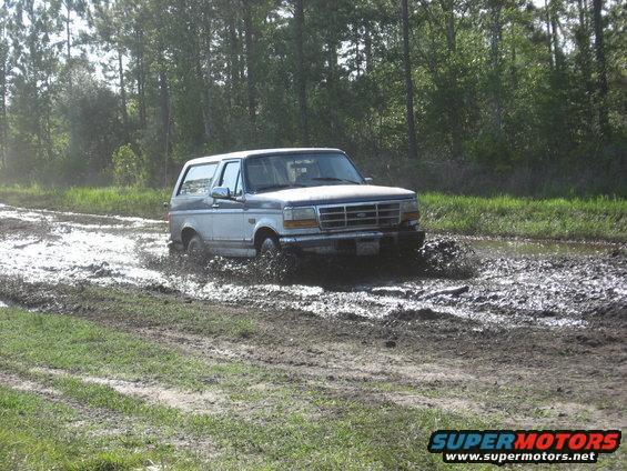 Two-tone blue and white Ford Bronco driving through mud, front 3/4 view